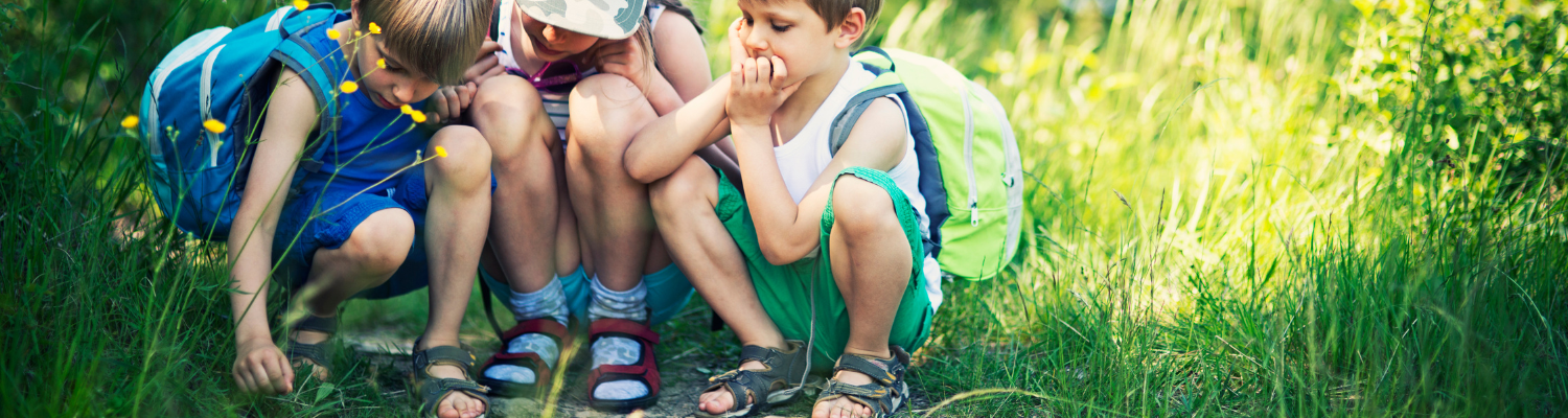 three children with packbacks looking at something in the grass