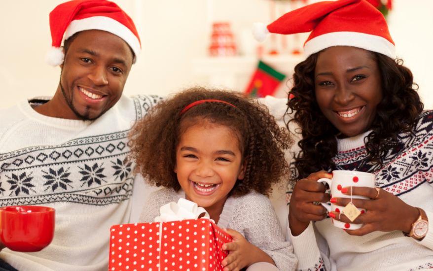 family of three sitting on couch with mugs and santa hats, christmas tree in background