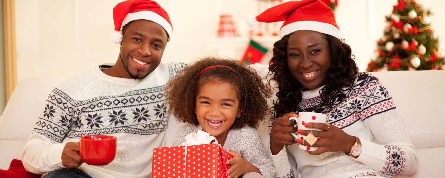 family of three sitting on couch with mugs and santa hats, christmas tree in background
